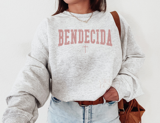 Light gray pullover sweater with the word "Bendecida" which means "Blessed" in english. There is a thin cross underneath the word, placed bottom center. Font resembles varsity letters in all caps. Font color is similar to a mauve blush, clay color, pink blush